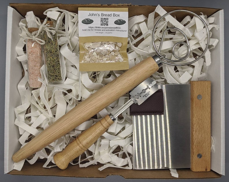 Puede incluir: Una caja blanca llena de papel de embalaje marr&oacute;n y una variedad de herramientas para hornear. Las herramientas incluyen un rodillo de madera, un raspador de masa, un batidor de masa, dos peque&ntilde;os frascos de vidrio y una peque&ntilde;a bolsa de papel marr&oacute;n con un c&oacute;digo QR. La bolsa tiene el texto "John's Bread Box" y una direcci&oacute;n de sitio web.