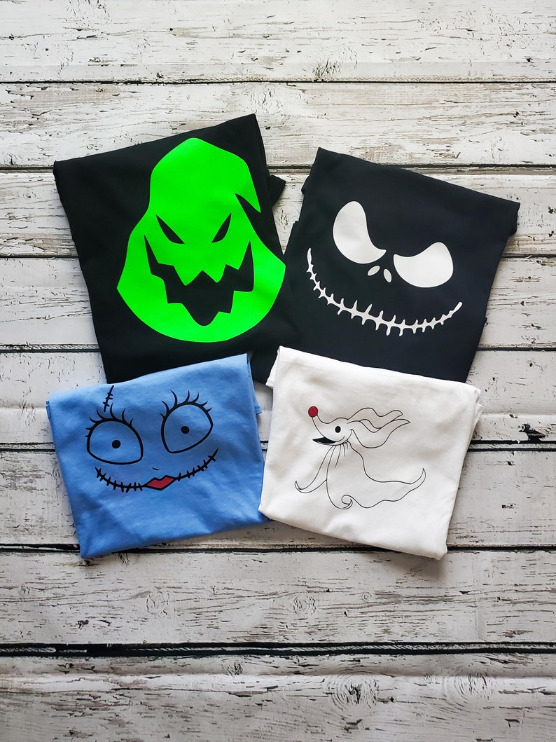 May include: Four folded t-shirts with different characters from the Nightmare Before Christmas movie. The shirts are black, white, blue, and green. The characters are Oogie Boogie, Jack Skellington, Sally, and Zero.