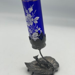 May include: A cobalt blue glass bud vase with a white floral design sits on a silver metal stand with a leaf and deer design.
