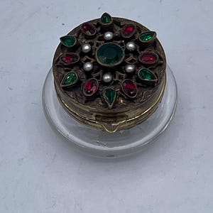 May include: A round, gold-colored trinket box with a glass base. The lid is decorated with a circular design of red and green gemstones and white pearls.
