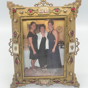 Könnte beinhalten: Verzierter goldfarbener Bilderrahmen mit einem Foto von drei Frauen. Der Rahmen hat rote und weiße Akzente, dekorative Schnörkel und kleine gemalte Szenen. Ein Rahmen im Vintage-Stil zum Präsentieren von Erinnerungen.