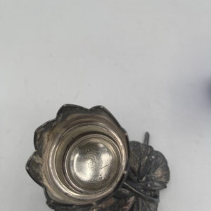 May include: Silver salt cellar with a leaf design base. The top is a round, silver cup with a decorative rim.