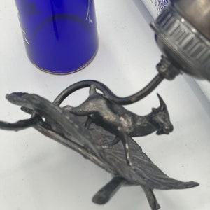 May include: A silver metal sculpture of a goat standing on a leaf-shaped base. The goat is facing to the right and has a long, curved horn. The base is made of metal and has a textured surface.