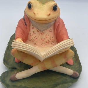 Könnte beinhalten: Eine Keramik-Froschfigur sitzt auf einem Seerosenblatt und liest ein Buch. Der Frosch trägt eine rosa Jacke und Weste, mit gekreuzten Beinen und braunen Schuhen. Die Haut des Frosches ist eine Mischung aus Grün und Gelb, mit großen schwarzen Augen.