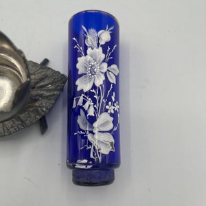 May include: A cobalt blue glass bud vase with a white floral design. The vase has a round base and a cylindrical shape.