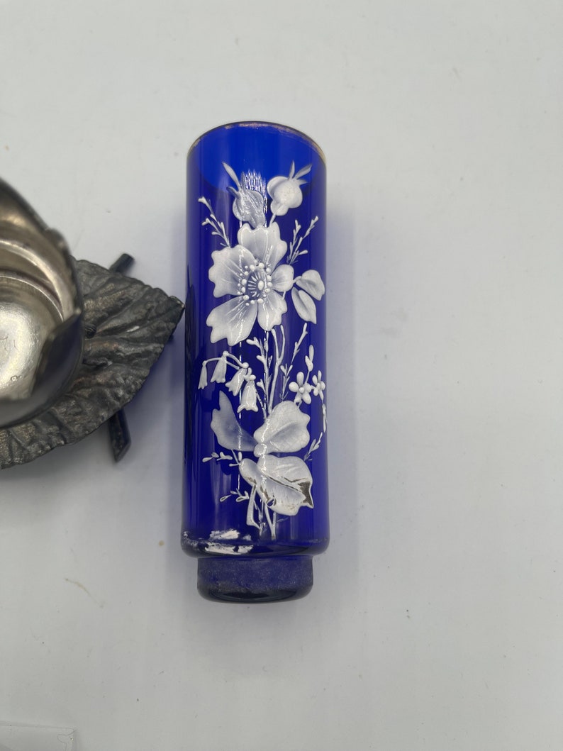 May include: A cobalt blue glass bud vase with a white floral design. The vase has a round base and a cylindrical shape.
