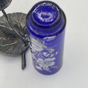 May include: A cobalt blue glass bottle with a white floral design. The bottle is topped with a silver metal lid and sits on a silver metal base.