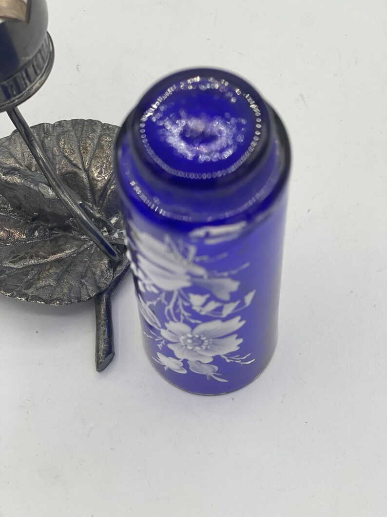 May include: A cobalt blue glass bottle with a white floral design. The bottle is topped with a silver metal lid and sits on a silver metal base.