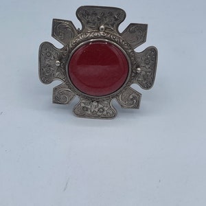 Op de afbeelding: Een zilveren broche met een rode steen in het midden. De broche heeft de vorm van een ster met ingewikkelde bloemmotieven.