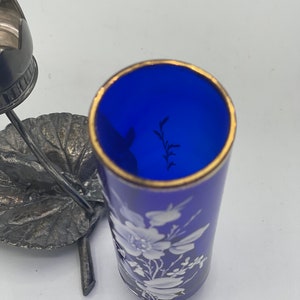 May include: A cobalt blue glass bottle with a gold rim and white floral design. The bottle is sitting on a silver leaf-shaped stand.