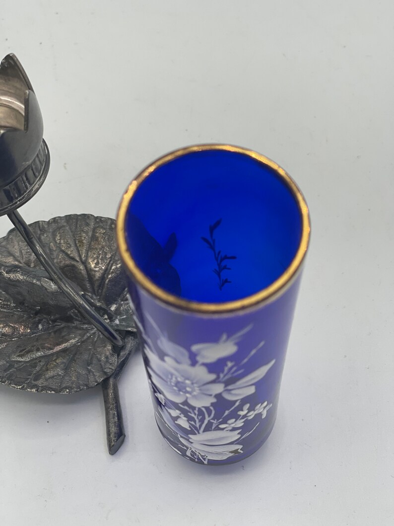 May include: A cobalt blue glass bottle with a gold rim and white floral design. The bottle is sitting on a silver leaf-shaped stand.
