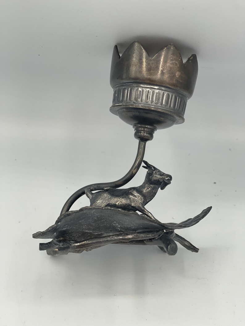 May include: Silver candlestick with a goat on a turtle base. The candlestick has a crown-shaped top.