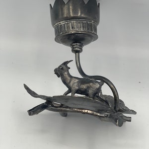 May include: Silver candlestick with a goat figurine standing on a leaf design base. The candlestick has a flower-shaped top.