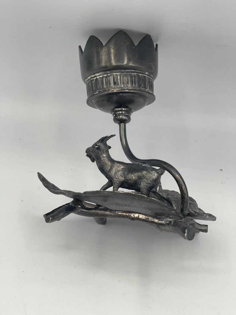 May include: Silver candlestick with a goat figurine standing on a leaf design base. The candlestick has a flower-shaped top.