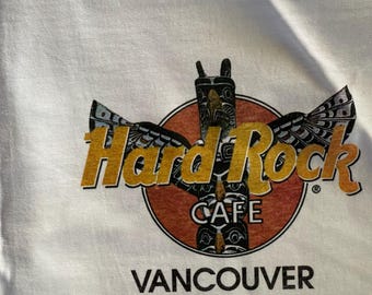 May include: White t-shirt with a Hard Rock Cafe logo featuring a totem pole and the words "Hard Rock Cafe Vancouver".