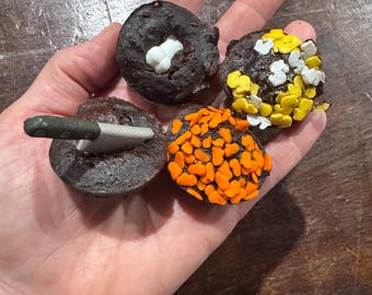 以下が含まれることがあります： ハロウィーンをテーマにしたトッピングで飾られた4つのミニチュアダークチョコレートマフィン。1つには小さなプラスチック製のナイフ、もう1つには白い骨、1つには黄色と白のトッピング、最後には鮮やかなオレンジ色のトッピングが施されています。