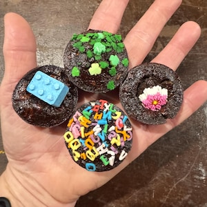 May include: Four chocolate cupcakes with various decorations. One has a blue Lego brick, another has green shamrock sprinkles, a third has a pink and white flower, and the last is covered in colorful alphabet sprinkles. The cupcakes are held in a hand.