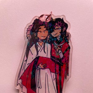 May include: A clear acrylic keychain featuring two anime characters, one with long black hair and a red and black outfit, and the other with long brown hair and a white outfit. The characters are holding each other and have a starry background.