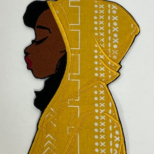 May include: A yellow embroidered iron-on patch featuring a profile of a woman with a hood. The hood has a white geometric pattern.