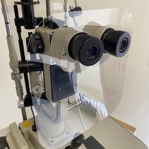 May include: A white and gray medical device with two black lenses. The device has a clear plastic shield in front of the lenses. The device has a black control panel with the text "SLIT LAMP" and "OLMPUS" printed on it.