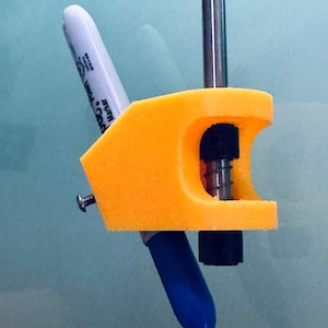 Op de afbeelding: Een gele 3D-geprinte houder met een zwarte schroef en een metalen staaf. Een blauwe en witte marker wordt in de houder gehouden.