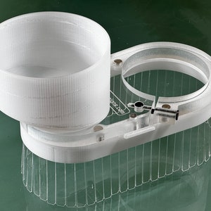 May include: A white 3D printed filament holder with a cylindrical container and a ring-shaped base with a textured edge. The holder has a small lever and a printed label that says "thingiverse.com".