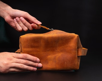 May include: A brown leather wash bag with a zip fastening and a handle. The bag is open and the zip pull is visible.