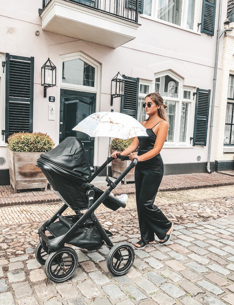 May include: A woman wearing sunglasses and a black jumpsuit pushes a black stroller with a white and gold umbrella. The stroller has a black leather seat and a black basket.
