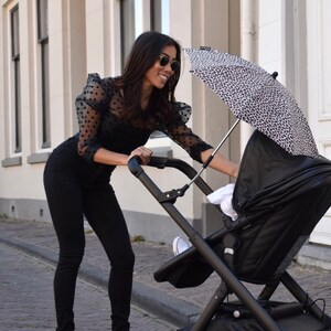 May include: A woman in a black polka dot blouse and black jeans pushes a black stroller with a black and white patterned umbrella attached. The stroller has a black canopy and a black basket underneath.