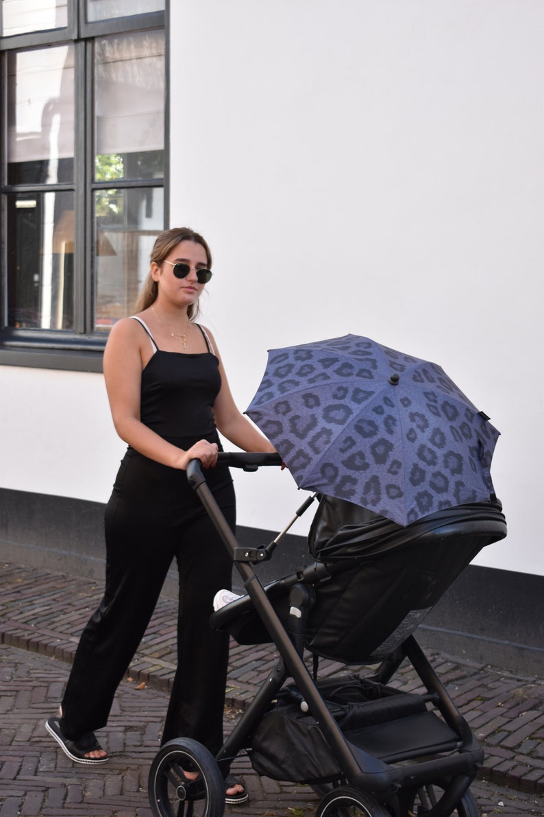 May include: A person pushing a black stroller with a black and blue leopard print umbrella attached. The person is wearing a black tank top and black pants.