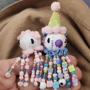 May include: Two small plush creatures with white fabric heads, black embroidered eyes, and pink pom-pom accents. The larger one wears a green star-patterned party hat. Both have dangling tentacles made of colorful plastic beads, including lettered cubes spelling DTC, dice, and eye-shaped beads.