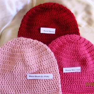 May include: Three crocheted beanies in shades of red and pink. The colors are labeled "Burnt Sienna", "Warm Blush (Lt. Pink)", and "Rosey/ Med. Pink". The beanies are displayed on a light-colored, textured surface.