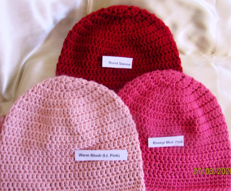 May include: Three crocheted beanies in shades of red and pink. The colors are labeled "Burnt Sienna", "Warm Blush (Lt. Pink)", and "Rosey/ Med. Pink". The beanies are displayed on a light-colored, textured surface.