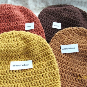 May include: Four crocheted beanies in various colors: spice, brown, antique gold, and mineral yellow. Each hat has a label with its color name. The hats are handmade and appear to be made of a soft yarn.