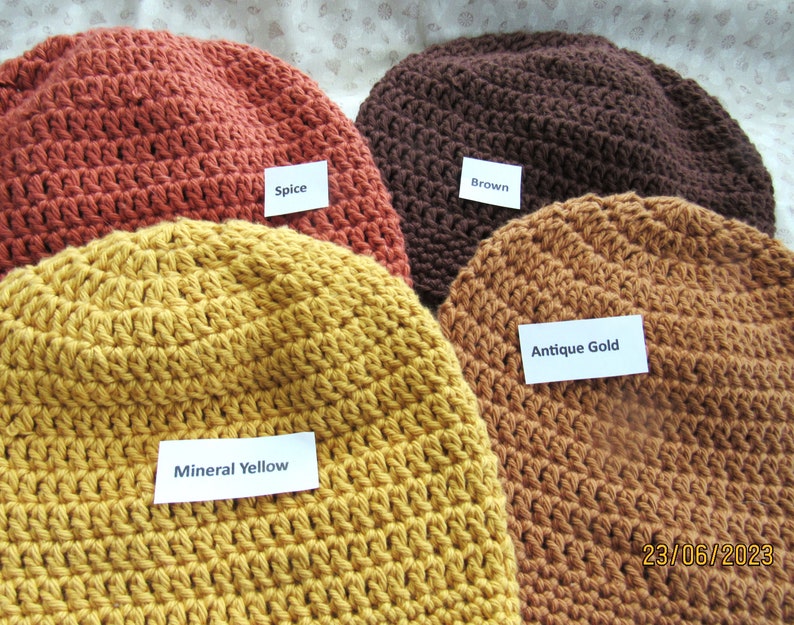 May include: Four crocheted beanies in various colors: spice, brown, antique gold, and mineral yellow. Each hat has a label with its color name. The hats are handmade and appear to be made of a soft yarn.