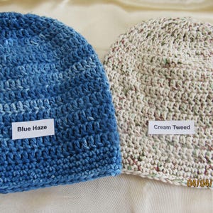 May include: Two crocheted beanies. One is blue with the text "Blue Haze" on a white label. The other is cream-colored with flecks of color and the text "Cream Tweed" on a white label. Both are handmade.