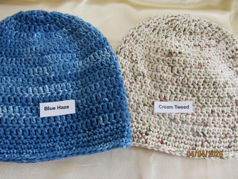 May include: Two crocheted beanies. One is blue with the text "Blue Haze" on a white label. The other is cream-colored with flecks of color and the text "Cream Tweed" on a white label. Both are handmade.