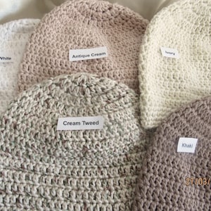 May include: A collection of handmade crochet beanies in various colors, including white, antique cream, ivory, cream tweed, and khaki. Each beanie has a label with its color name. The beanies are made of a textured yarn.