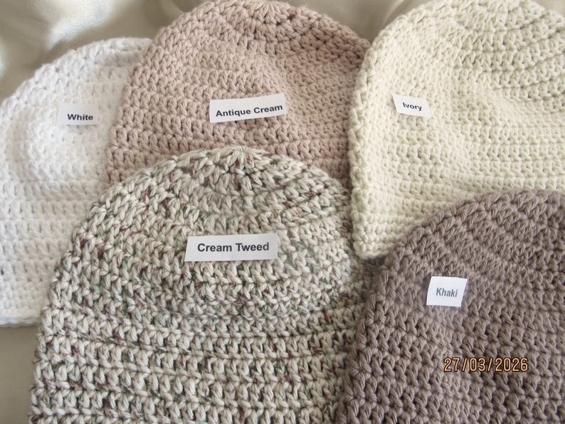 May include: A collection of handmade crochet beanies in various colors, including white, antique cream, ivory, cream tweed, and khaki. Each beanie has a label with its color name. The beanies are made of a textured yarn.