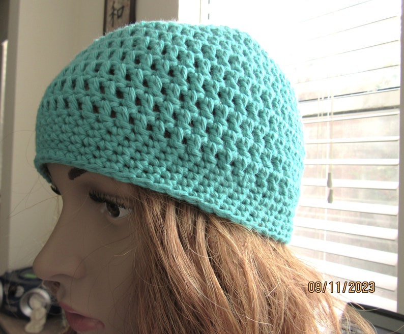 May include: A turquoise crochet beanie hat. The hat is made with a textured stitch pattern, creating a breathable design. The hat is a solid color and appears to be handmade. The beanie is a close-fitting style, suitable for casual wear.