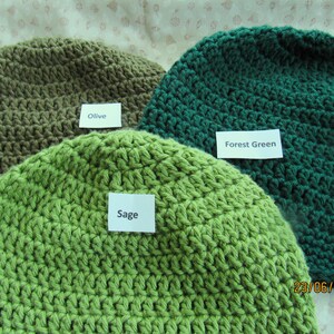 May include: Three crocheted beanies in shades of green and olive. The beanies are labeled "Olive", "Sage", and "Forest Green". The hats are handmade with a visible stitch pattern. The date "23/06/2023" is visible in the bottom right corner.