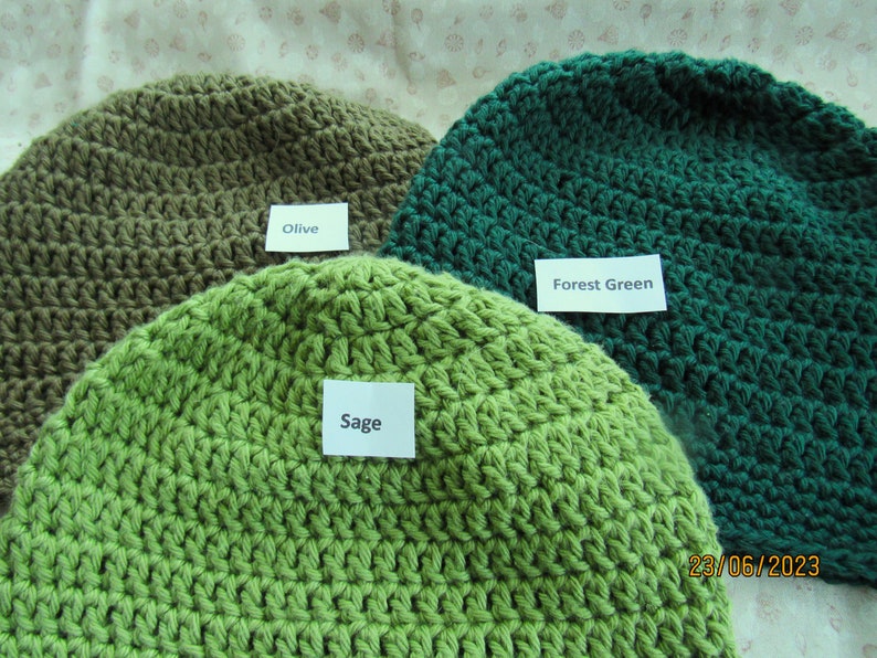May include: Three crocheted beanies in shades of green and olive. The beanies are labeled "Olive", "Sage", and "Forest Green". The hats are handmade with a visible stitch pattern. The date "23/06/2023" is visible in the bottom right corner.