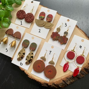 Könnte beinhalten: Sortiment von 10 Paar handgefertigten Polymer Clay Ohrringen mit goldenen Akzenten. Die Ohrringe haben verschiedene Formen, darunter Kreise, Blätter, Blumen und geometrische Muster. Die Ohrringe werden auf weißen Karten mit den Nummern 1 bis 7 angezeigt.