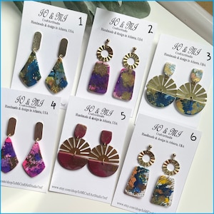 May include: Six pairs of handmade earrings with different designs and colors. The earrings are made with resin and have a variety of shapes, including circles, triangles, and sunbursts. The earrings are all gold-toned and have a unique, artistic look.