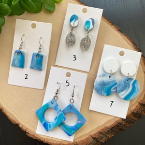 Könnte beinhalten: Fünf Paar blaue und weiße Polymer Clay Ohrringe, die auf weißen Karten mit den Nummern 2, 3, 5 und 7 präsentiert werden. Die Ohrringe haben verschiedene Formen, darunter Tropfen, Quadrat, Kreis und Diamant.