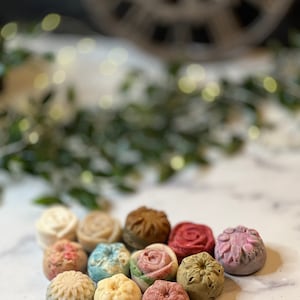 May include: Assortment of small, round, floral-shaped soaps in various colours, including pink, green, blue, white, and brown. The soaps are arranged on a white marble surface.