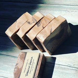 May include: Five bars of handmade soap with a tobacco vanilla scent. The bars have a marbled design with shades of brown and tan. One bar is wrapped with a label that reads "Tobacco Vanilla" and "Net Wt. 4.5 oz."