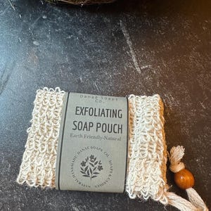 Puede incluir: Una bolsa de jabón exfoliante de color natural con una etiqueta que dice "EXFOLIATING SOAP POUCH" y "Earth Friendly-Natural". La bolsa tiene un lazo con una cuenta de madera. El producto es de Danae Soaps Co.
