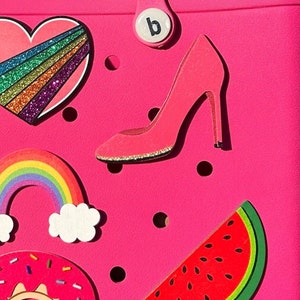 May include: A bright pink Bogg tote bag featuring two handles and the word bogg embossed on the front. The bag is decorated with various colorful charms including a green frog, a glittery rainbow heart, a pink high heel, a rainbow, a lemon, a donut, and a watermelon slice.