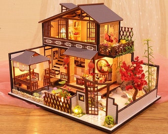May include: A miniature wooden dollhouse with a traditional Japanese design. The house has a two-story structure with a balcony, a red roof, and a small garden with a pond and a red tree. The house is lit up with warm lights.
